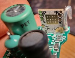 A circuit board with a chip against a finger, showing board markings and a PET capacitor.