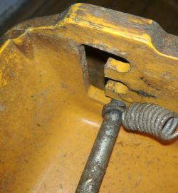 Zbliżenie na żółtą obudowę kosiarki z widoczną sprężyną i metalową osią, ukazujące mechanizm regulacji wysokości.