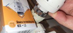 Ręka trzymająca głowicę termostatyczną nad kaloryferem z opakowaniem w tle.