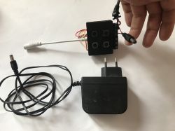 A hand holds a DC plug connected to a black module with several buttons. Next to it lies a 12V power adapter with a cord and DC plug.