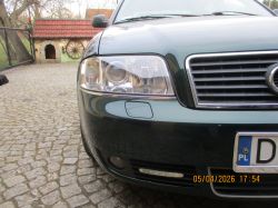 Przód zielonego auta z reflektorem i grillem na brukowanym podjeździe, w tle budynek z czerwonym dachem