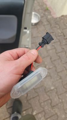 Dłoń trzyma lampkę LED do podświetlenia tablicy z wtyczką i przewodami czerwonym i czarnym.