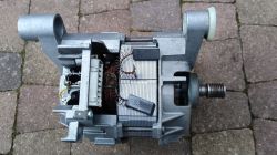 Silnik elektryczny wyjęty z obudowy, z widocznymi szczotkami i uzwojeniami na tle brukowanej powierzchni.