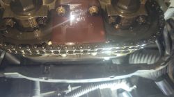 Zbliżenie na łańcuch rozrządu oraz napinacz w silniku Opel Corsa A12XER z widocznym olejem silnikowym.