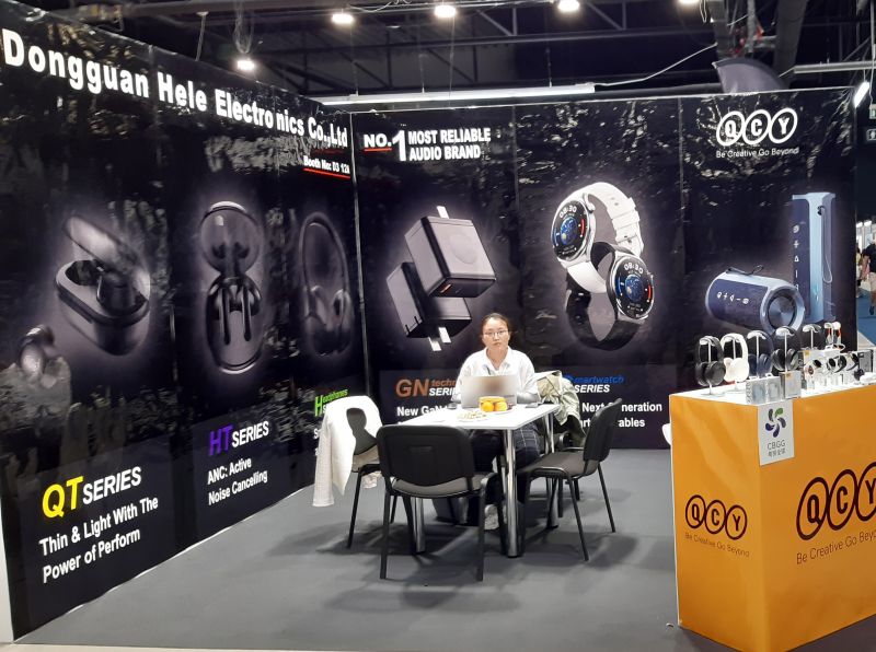 Stoisko firmy Dongguan Hele Electronics Co., Ltd na targach elektroniki z plakatami reklamującymi słuchawki i smartwatche.