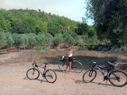 Osoba stoi obok roweru na tle górzystego krajobrazu z drzewami. Obok znajdują się jeszcze dwa rowery.