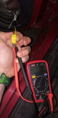 Osoba trzyma żółtą wtyczkę i mierzy napięcie miernikiem samochodowym.