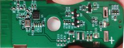 Close-up of a green PCB with SMD components, a mounting hole, and a plus sign marking