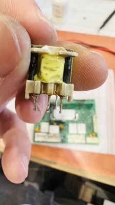 Zbliżenie na transformator trzymany w dłoni przed płytką elektroniczną.
