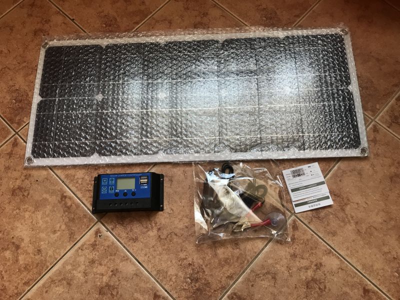 Solar kit: photovoltaic panel in bubble wrap, charge controller, accessories, and manual on the floor.