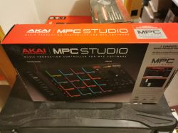 Pudełko z kontrolerem muzycznym AKAI Professional MPC Studio, widoczne na półce wśród innych sprzętów.