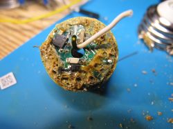 Close-up of a damaged and corroded electronic circuit board with visible rust, debris, and a protruding white wire.