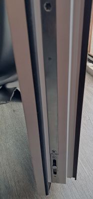 Widok mechanizmu zamka w uchylonym skrzydle drzwiowym z widocznymi śrubami i prowadnicą.
