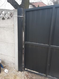 Uszkodzony zawias bramy zamocowany do słupka, brama wisi na jednym zawiasie.