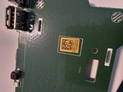 Close-up of a green PCB with a USB port, a small button, and a yellow label with a 2D code.