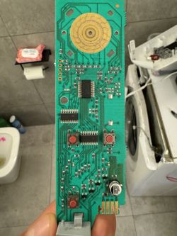 Płytka drukowana z elementami elektronicznymi trzymana w ręku, w tle pralka i toaleta.
