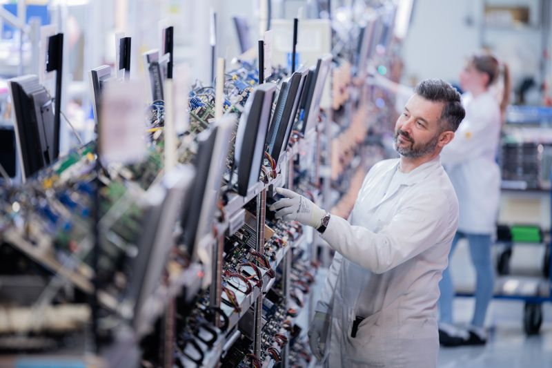 Mitarbeiter im weißen Kittel prüft Elektronik an einer Montagelinie mit Monitoren
