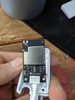 Circuit board with a metal shield and white connector, held in a hand above a wooden surface
