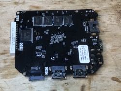 Czarna płytka PCB od przystawki TV Box z widocznymi układami scalonymi, złączami USB i Ethernet, leżąca na drewnianej powierzchni.