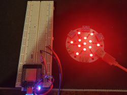 Breadboard with a microcontroller module and wires next to a round LED board glowing red