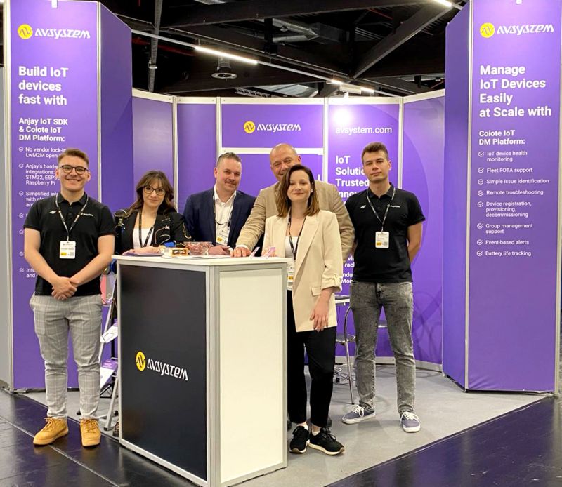Stoisko firmy AVSystem na targach, z sześcioma osobami stojącymi przed ladą i fioletowymi banerami w tle.