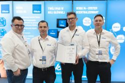 Four men in white shirts at a trade booth; one holds an open diploma, blue promotional panels in the background.