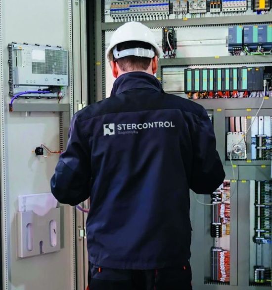 Technik w kasku przed otwartą szafą sterowniczą z modułami PLC i okablowaniem