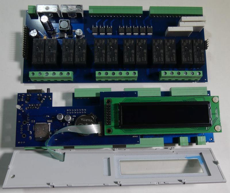 Dwie niebieskie płytki PCB z przekaźnikami i złączami oraz moduł LCD; na dole szara obudowa z okienkiem.