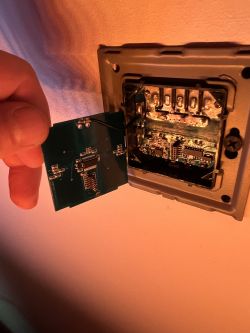 Person holding an electronic circuit board next to a wall-mounted panel.