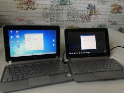 Dwa laptopy HP Mini 210 stojące obok siebie na tle ściany z graffiti, na obu widoczne ekrany ze szczegółami sprzętowymi.