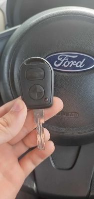 Na zdjęciu widoczny jest kluczyk z pilotem samochodowym trzymany w ręce, na tle kierownicy z logo Ford.