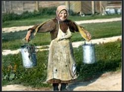A woman in a headscarf and apron carries two metal buckets outside.
