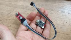 Dwie płytki elektroniczne i przewód na dłoni, jedna z nich to konwerter USB-UART, druga to moduł z przetwornicą.