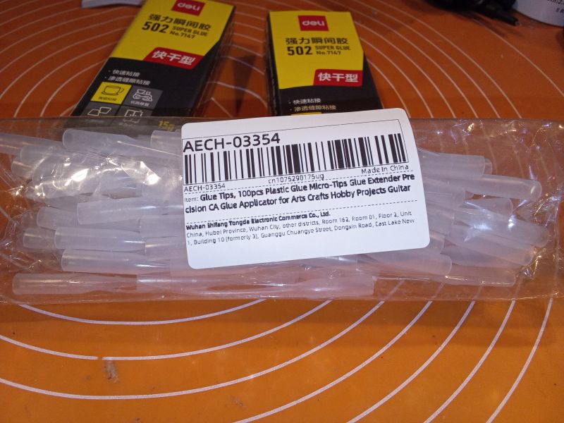 Opakowanie z plastikowymi końcówkami do precyzyjnego nakładania kleju oraz pudełka z klejem 502 na pomarańczowym tle.