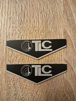 Dwa metalowe emblematy z logo TLC na drewnianej powierzchni.