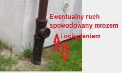 Rura spustowa przy ścianie budynku z czerwonymi strzałkami i napisem informacyjnym