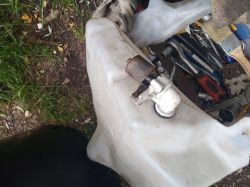 Zbiornik spryskiwaczy z zamontowanym silnikiem zapasowym, widoczny na tle narzędzi i trawy.