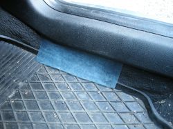 Kawałek folii wciśnięty w szczelinę między progiem drzwi a gumową wycieraczką w samochodzie.