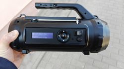 Black portable speaker with handle, antenna, and radio display, held in a hand outdoors