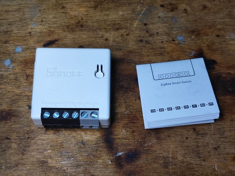 Sonoff ZBMini Relay with Manual Small Sonoff ZBMini relay next to a user manual on a wooden surface.