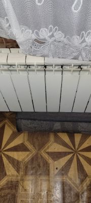 Aluminiowy grzejnik z zasłoną powyżej, na podłodze leży dywanik.