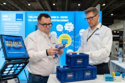 Two men in “Eltako” shirts at a trade booth reading a brochure beside blue Eltako crates.