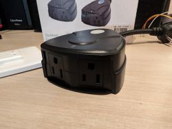 Prime Smart Outdoor Outlet with Remote Control Black smart outlet with two plugs on a wooden desk, with a printed package graphic of the device in the background.