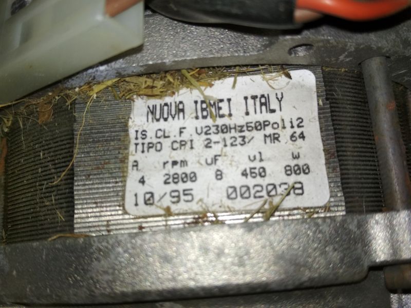 Tabliczka znamionowa silnika elektrycznego kosiarki, widoczne dane techniczne i zanieczyszczenia trawą.
