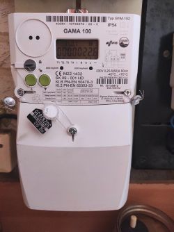 Elektroniczny licznik energii elektrycznej GAMA 100 z plombą zabezpieczającą zamontowany na ścianie.
