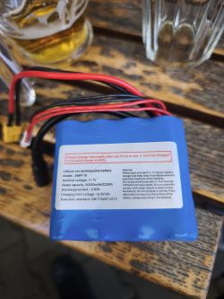Niebieska bateria Li-ion 11,1 V z przewodami, leżąca na drewnianym stole obok szklanek