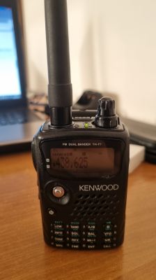 Ręczny radiotelefon Kenwood TH‑F7 z anteną, stojący na biurku obok laptopa.