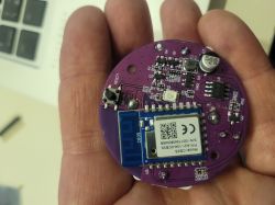 A person holds a round purple circuit board with various electronic components, including a wireless module.