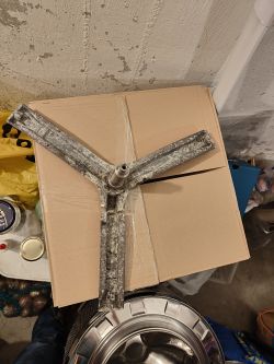 Metalowy krzyżak bębna pralki leży na kartonowym pudełku w pomieszczeniu gospodarczym.