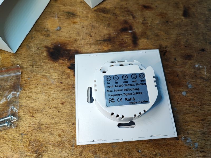 Smart Zigbee Light Switch View of the back of a white Zigbee-compatible light switch in packaging, on a wooden surface, next to a pack of screws.